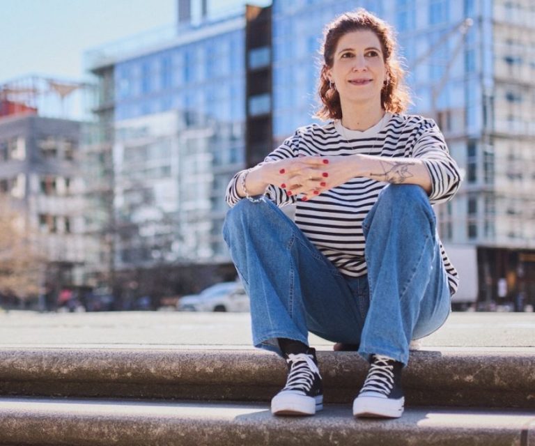 Das Bild zeigt Katharina Mandlinger, auf einer steinernen Stufe sitzend. Im Hintergrund zu sehen sind moderne Gebäude mit Glasfassaden.