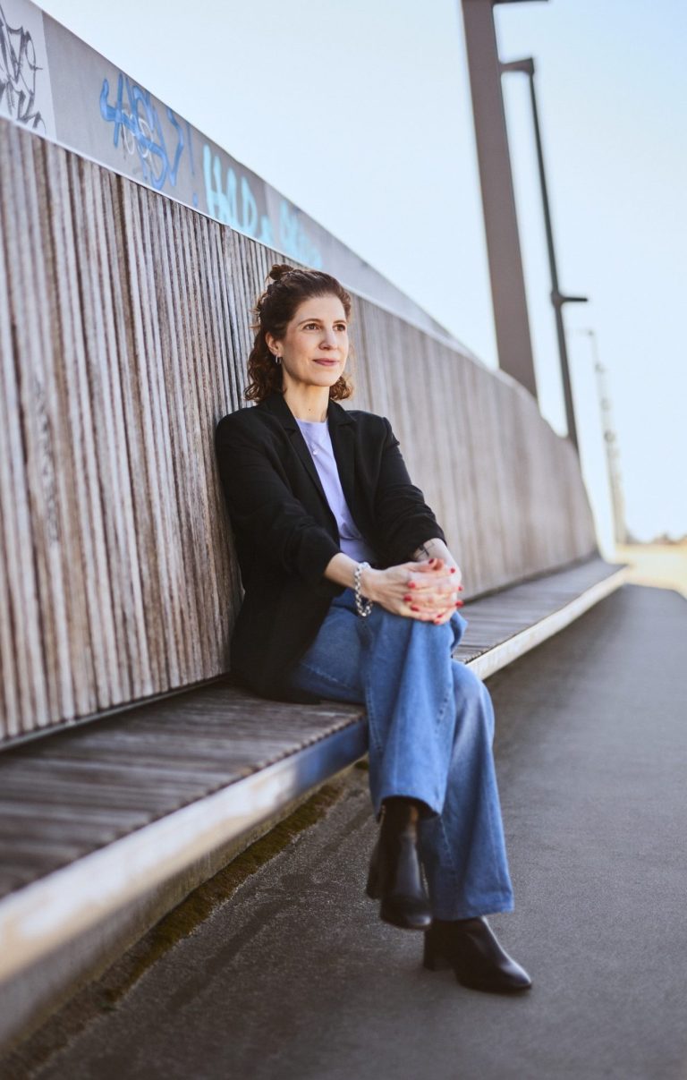 Das Bild zeigt Katharina Mandlinger, mit übereinandergeschlagenen Beinen sitzend auf einer langen Bank in der Hamburger HafenCity.